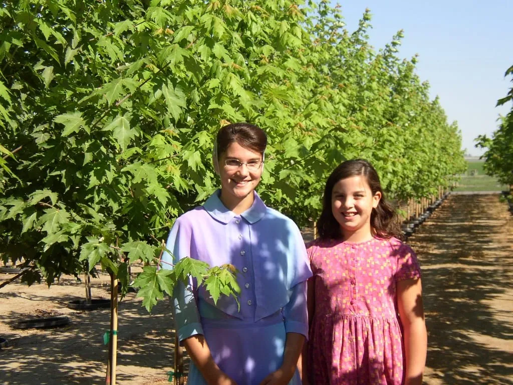 Two girls smiling between rows of young maple trees in pot-in-pot containers