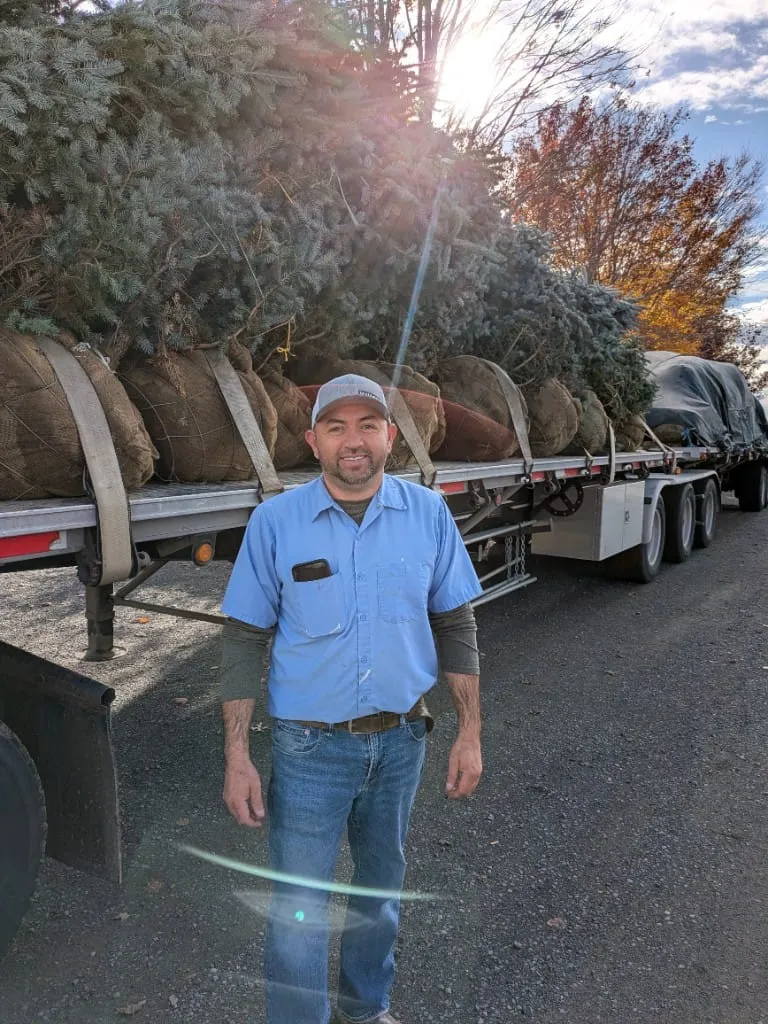 Ismael smiling in front of a flatbed trailer loaded with balled evergreen trees